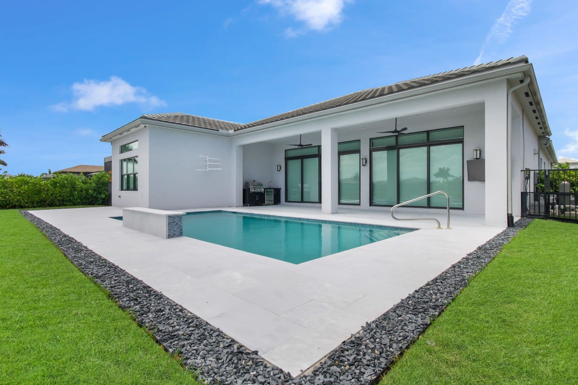 Modern white home with pool featuring large glass sliding doors, concrete patio, turquoise pool with handrail, manicured lawn, and metal roof with overhanging eaves
