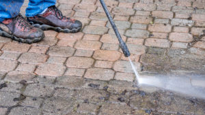 Person wearing jeans and work boots is cleaning paved driveway with pressure washer.